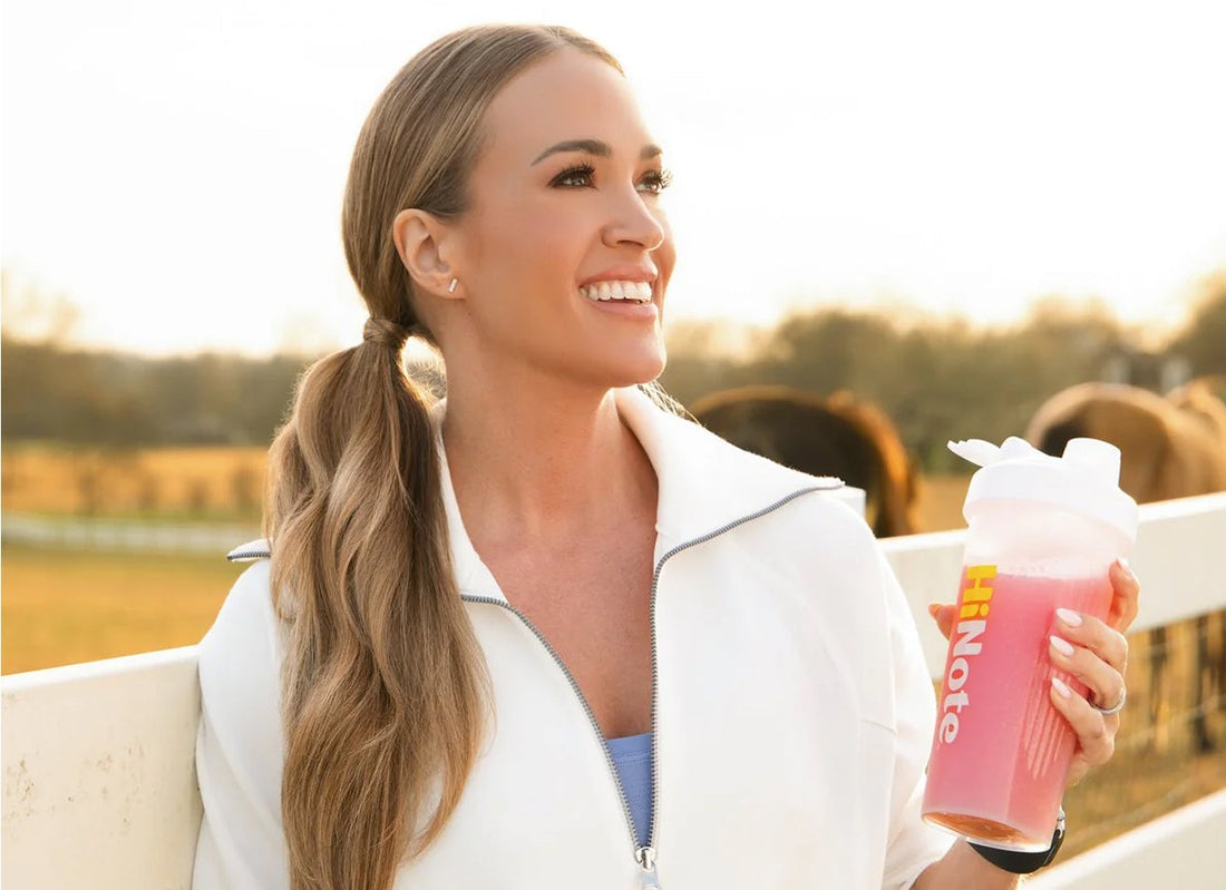 Carrie Underwood holding a pink HiNote shaker by a white fence at sunset with horses in the background