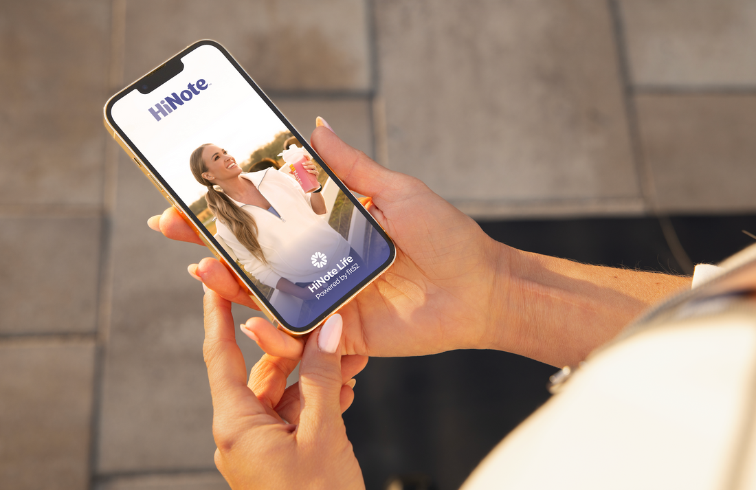 Person holding smartphone showing HiNote app splash reading HiNote and HiNote Life Powered by fit52 over outdoor tiles