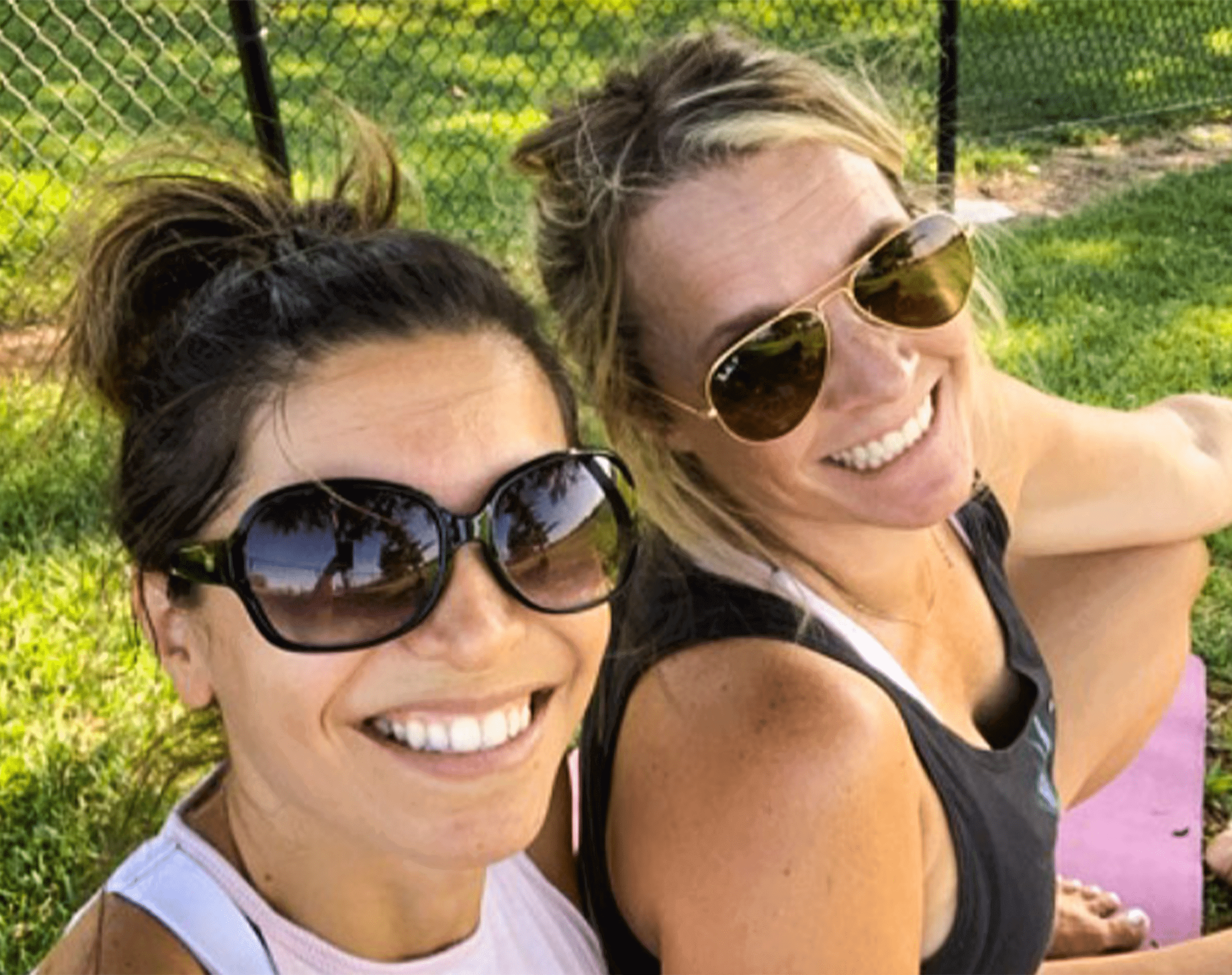 Two women wearing sunglasses smiling for a selfie on grass near a chain link fence