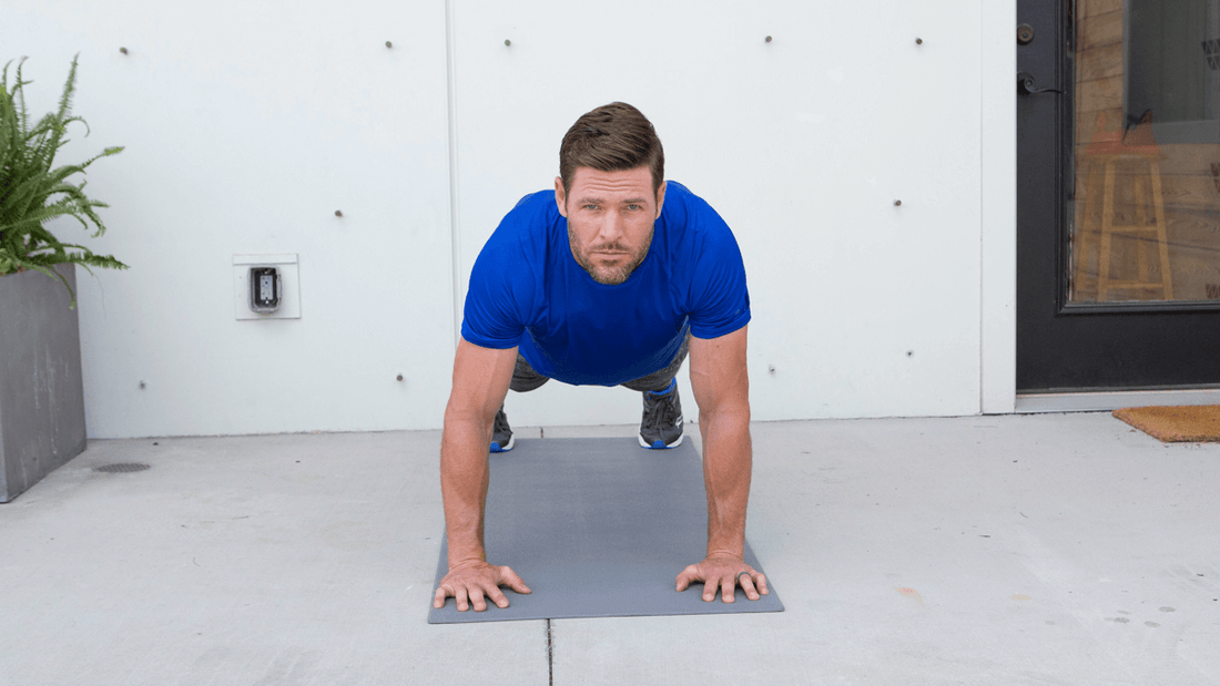 Man in a blue shirt holding a plank on a gray exercise mat outside a modern building entrance