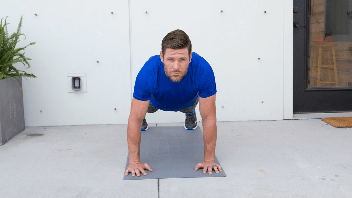 Man in a blue shirt holding a plank on a gray exercise mat outside a modern building entrance