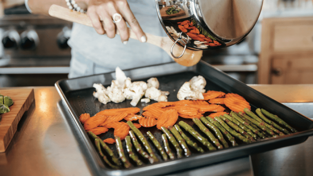 Hands pouring seasoned asparagus, crinkle-cut carrots, and cauliflower from a saucepan onto a baking sheet in a kitchen
