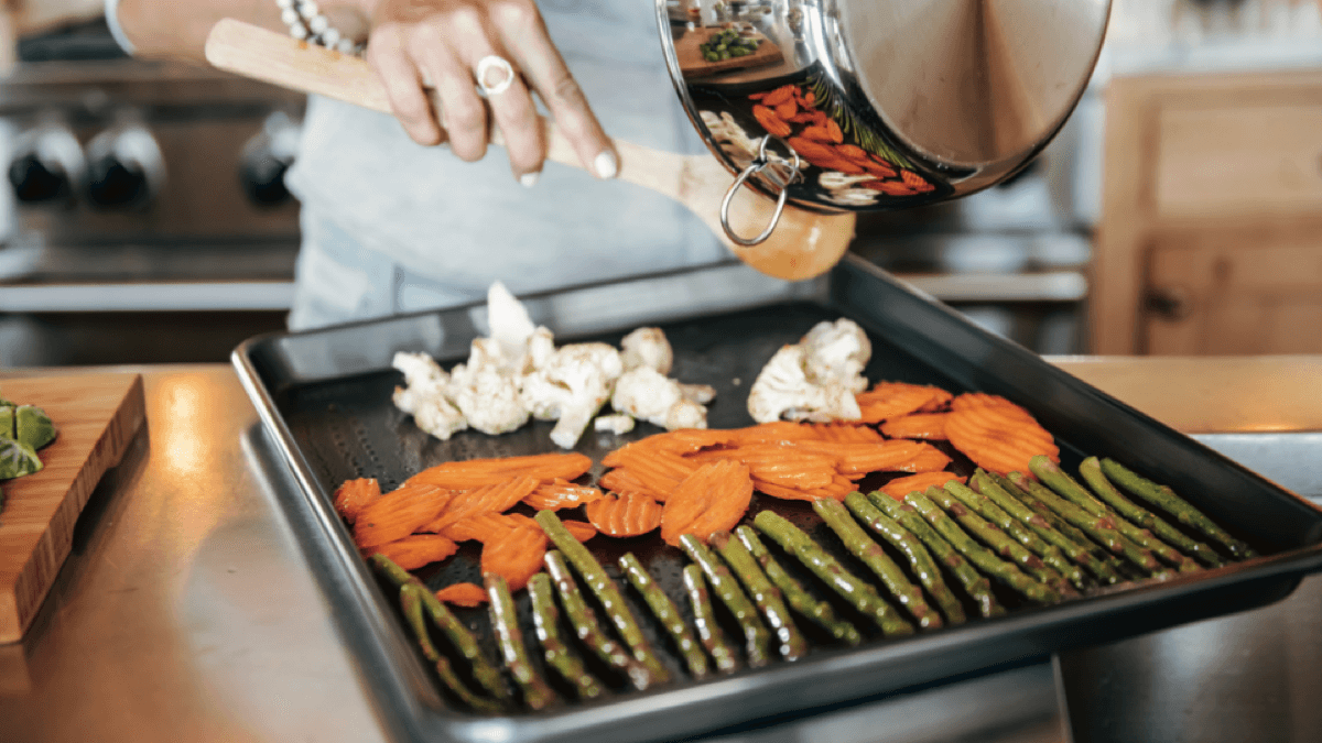 Hands pouring seasoned asparagus, crinkle-cut carrots, and cauliflower from a saucepan onto a baking sheet in a kitchen