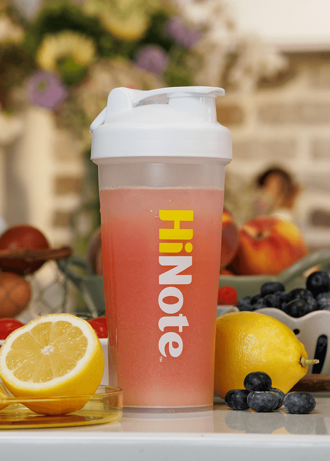 Front view of HiNote shaker bottle filled with pink drink, white flip lid and vertical HiNote logo, with lemons and blueberries on a blurred kitchen counter