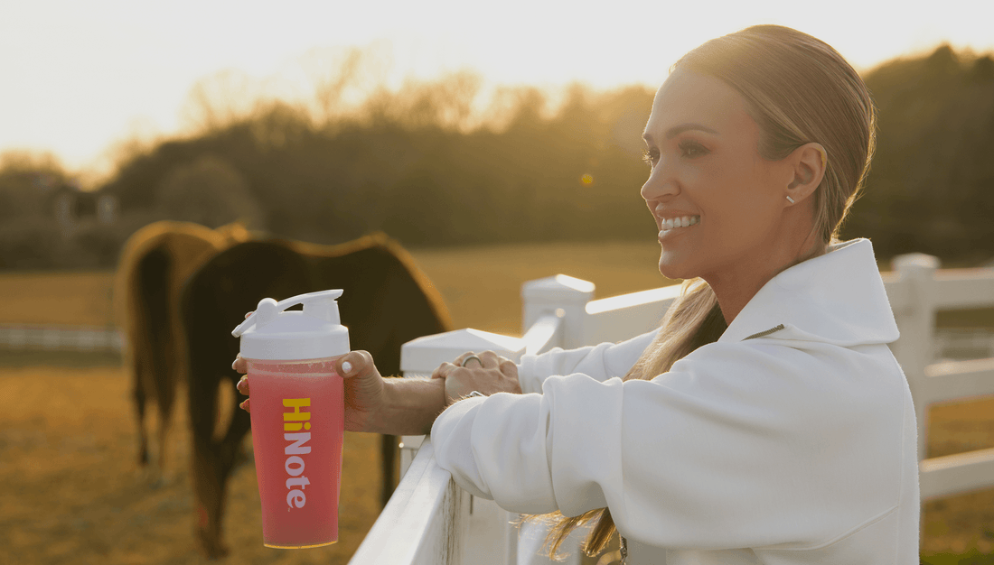 Carrie Underwood smiling at sunset leaning on a white fence while holding a pink HiNote shaker cup with horses grazing in the background