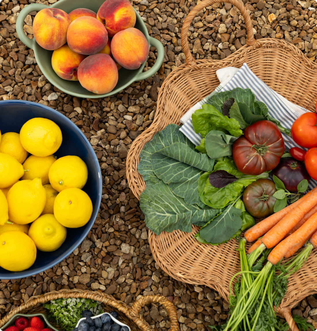 Fresh produce arranged in baskets on gravel, featuring lemons, peaches, carrots, leafy greens, tomatoes, and berries