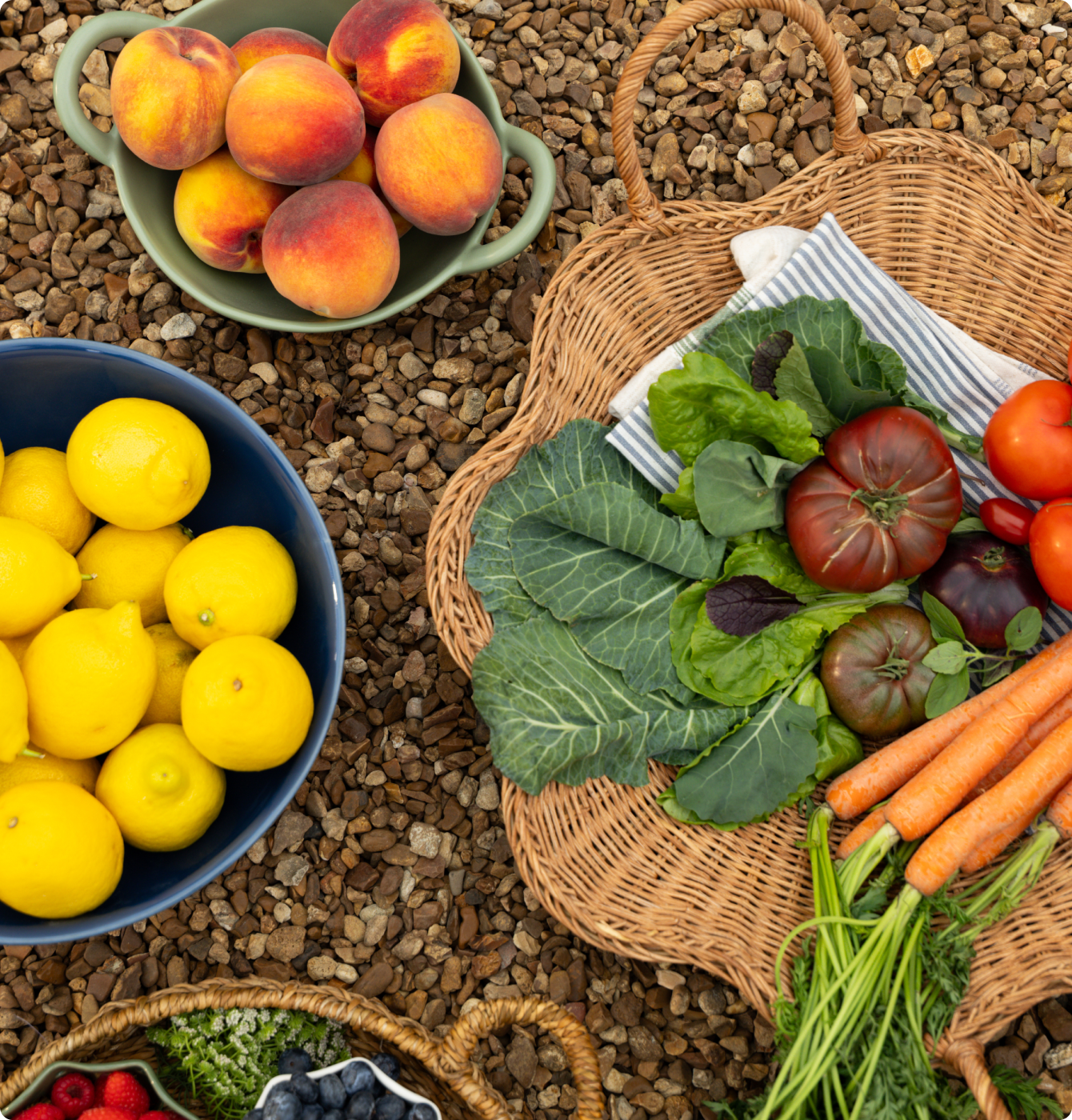 Fresh produce arranged in baskets on gravel, featuring lemons, peaches, carrots, leafy greens, tomatoes, and berries