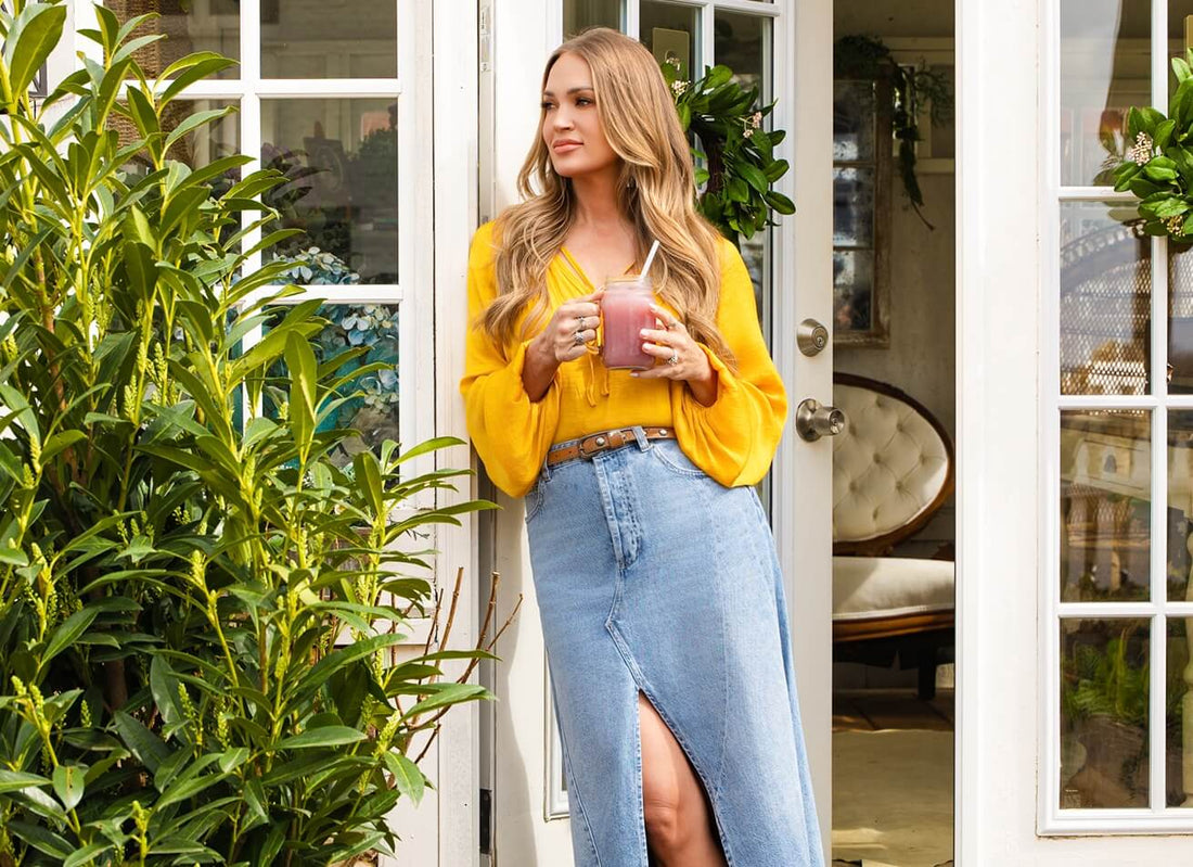 Carrie Underwood leaning in a sunlit doorway holding a pink smoothie in a jar, wearing a yellow blouse and denim skirt