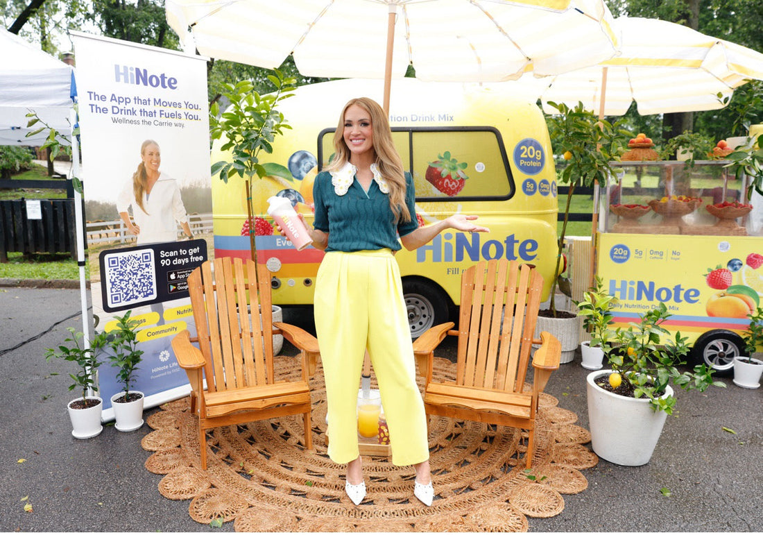 Carrie Underwood standing and smiling at an outdoor HiNote promo booth, holding a pink HiNote shaker in front of a branded van and banner