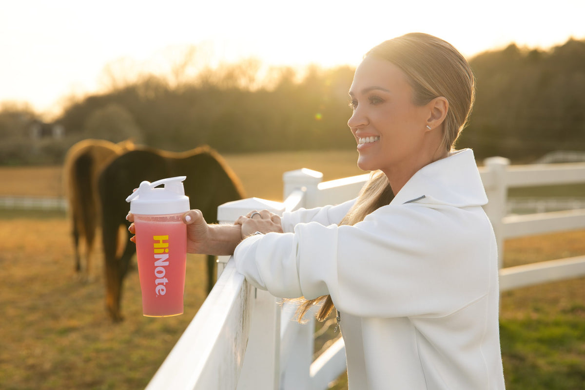 Carrie Underwood holding a pink HiNote bottle by a fence with a horse at sunset
