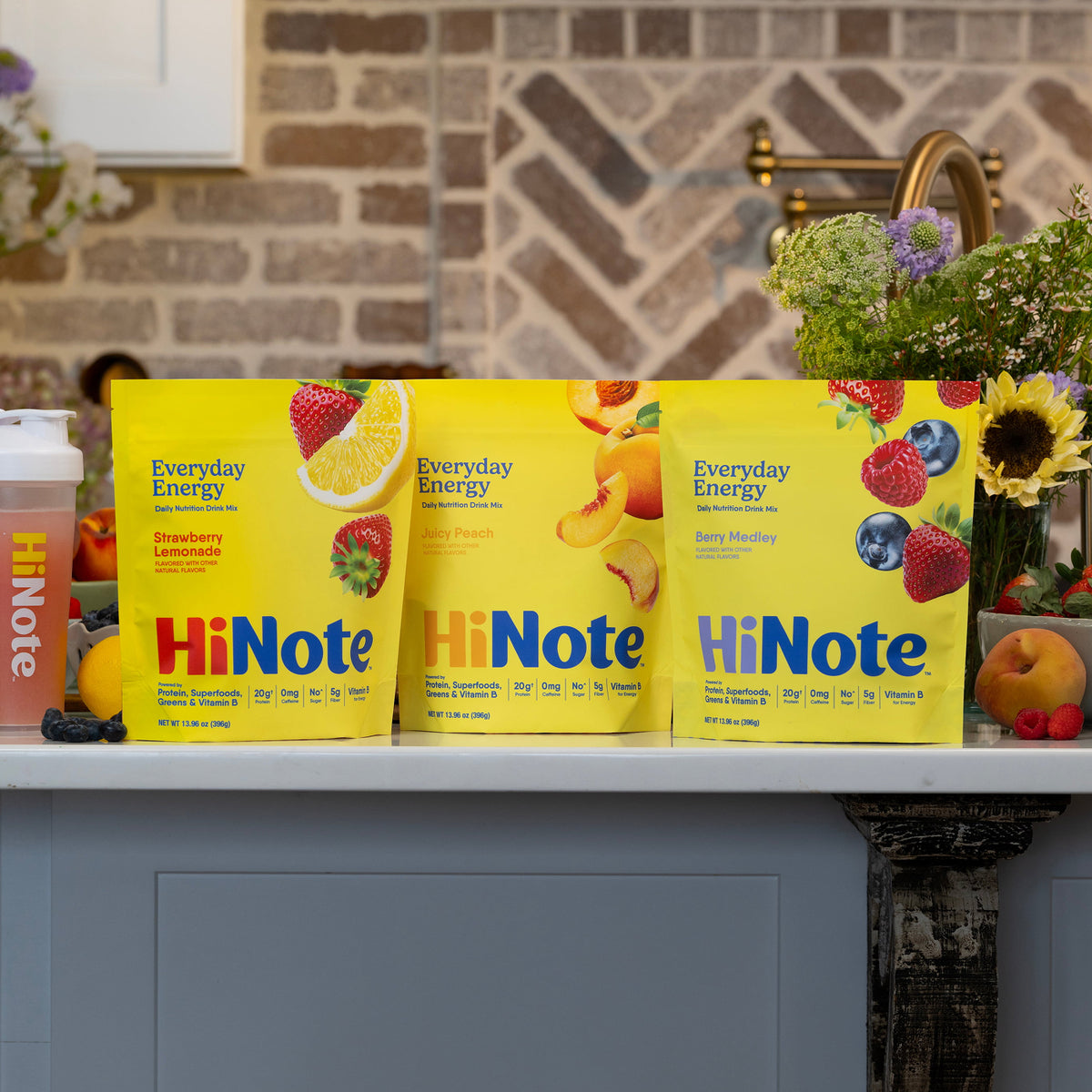 Three HiNote Everyday Energy bags in Strawberry Lemonade, Juicy Peach and Berry Medley on a kitchen counter next to a HiNote shaker bottle, fresh fruit and flowers