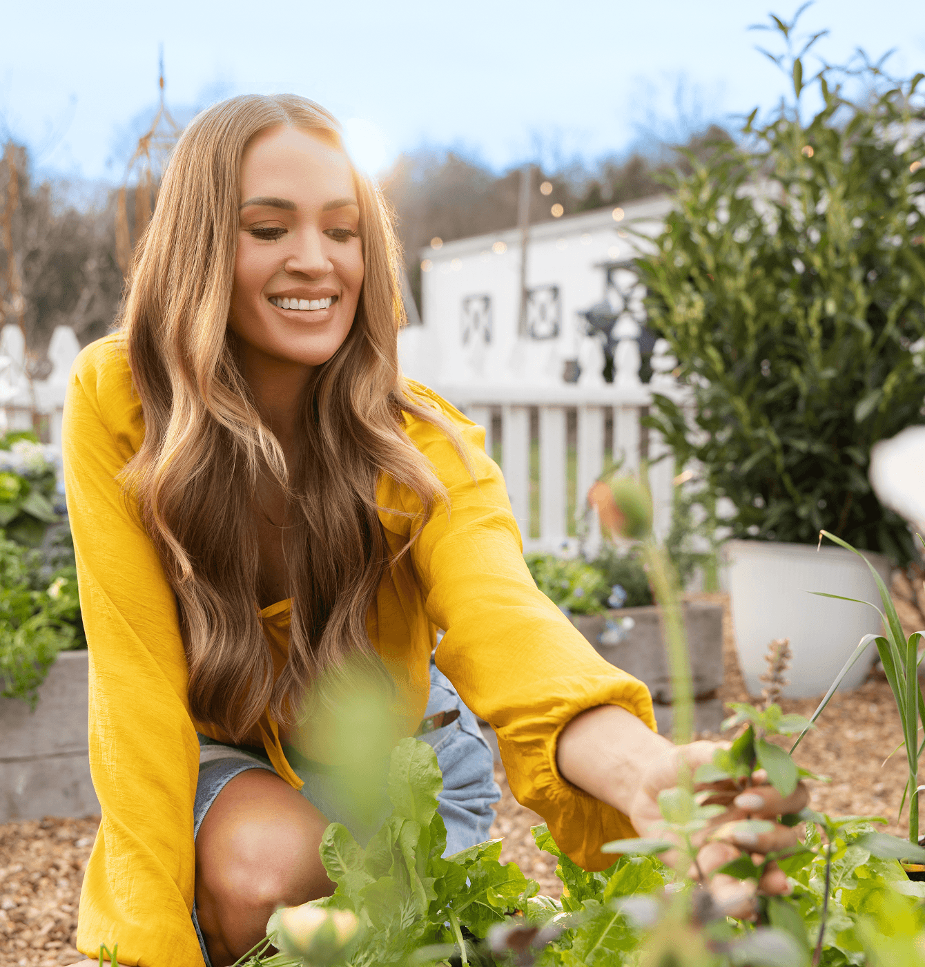 Carrie Underwood smiling while kneeling in a raised garden bed tending leafy greens with a white picket fence and string lights behind her