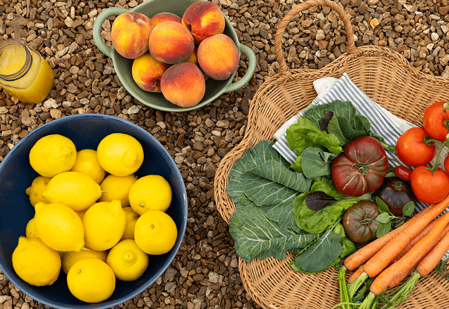 Bowl of lemons, basket of vegetables, and bowl of peaches on a stone surface