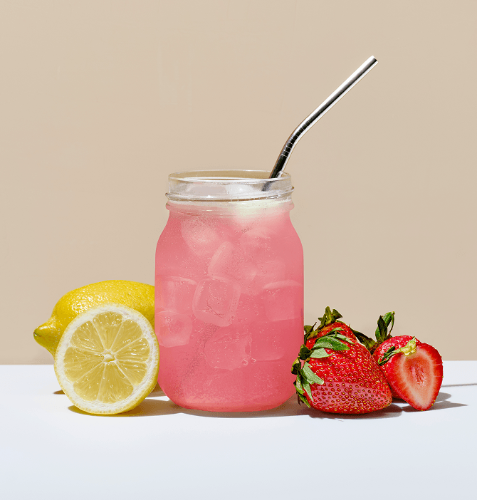 Front view of strawberry lemonade in a mason jar showcasing ice and a metal straw with lemon half and strawberries on a white tabletop