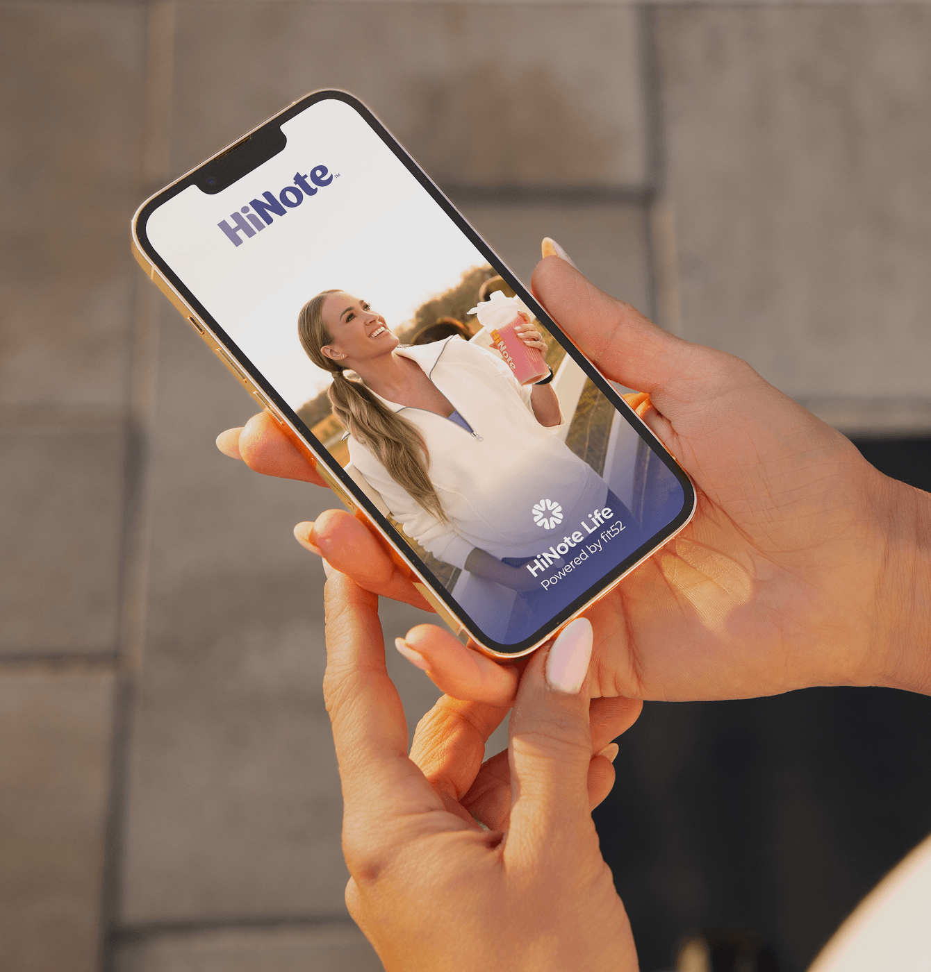 Angled smartphone view of HiNote Life app showing HiNote logo and smiling Carrie Underwood held in hands over blurred pavement