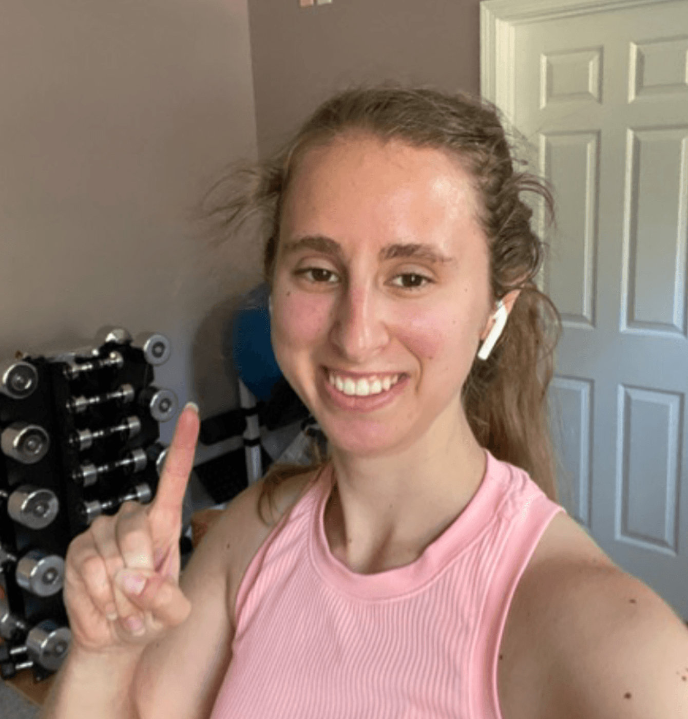 Woman smiling and pointing up while wearing wireless earbuds in a home workout room with a rack of dumbbells