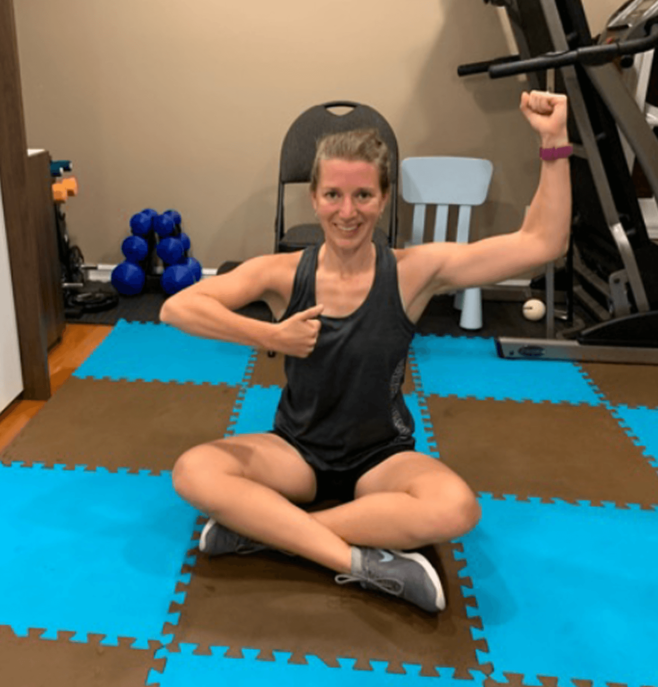 Woman sitting cross-legged on foam mats, flexing one arm and pointing to her chest in a home workout space with dumbbells and a treadmill