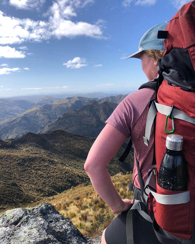 Woman with red backpack and water bottle standing on rocky ridge, looking over a sunlit mountain range under a blue sky