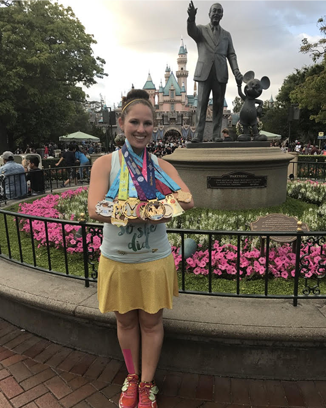 Woman smiling and showing multiple race medals while standing in front of the Partners statue and castle at a theme park