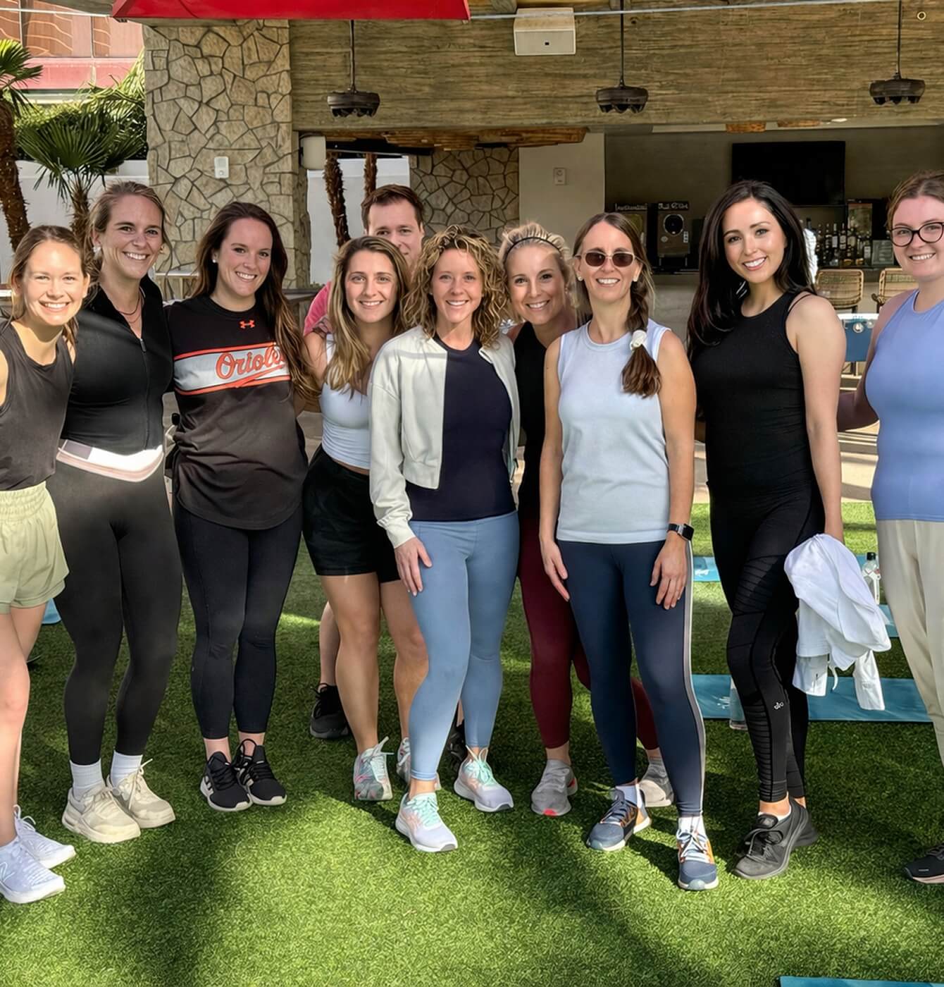 Group of women posing and smiling together outdoors on green turf after a workout, wearing casual activewear and sneakers