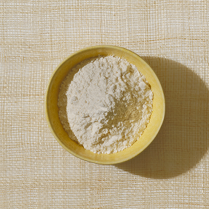 Top-down view of clear whey protein powder in a yellow bowl showcasing fine pale powder texture on a woven beige background