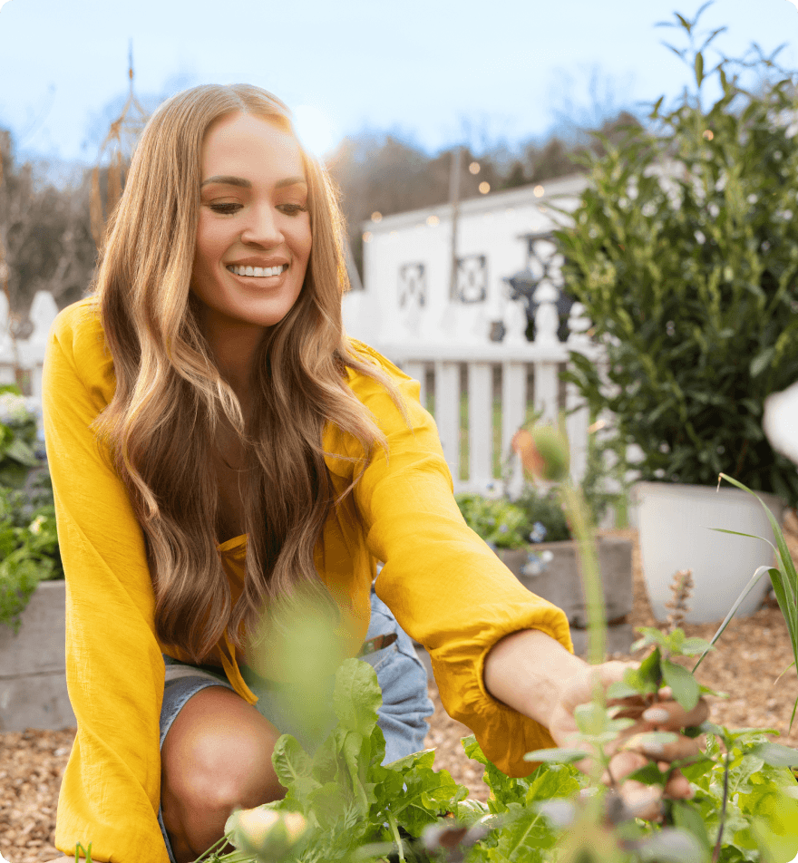 Carrie Underwood kneeling in a sunny backyard garden tending leafy greens, smiling with potted plants and a white fence behind her