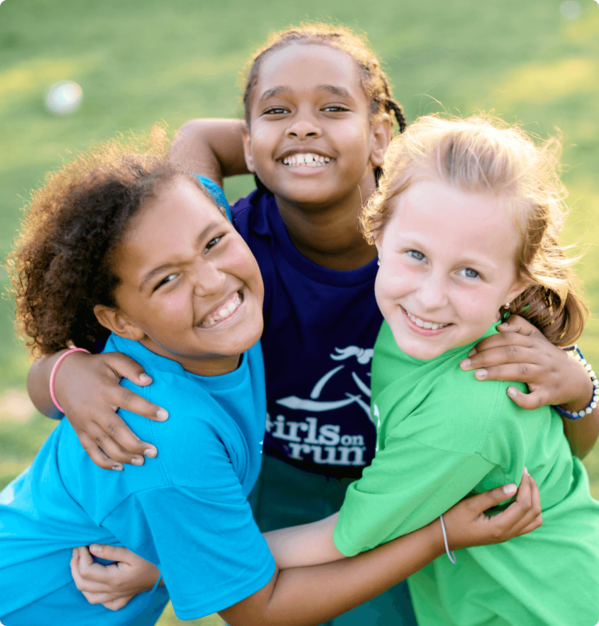 Three girls smiling and hugging on a grassy field at a Girls on the Run event