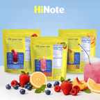 Three HiNote yellow drink-mix pouches, back panels showing the headline 'Fill your cup' and nutrition facts, with strawberries, blueberries, citrus slices, and a glass of pink drink in front
